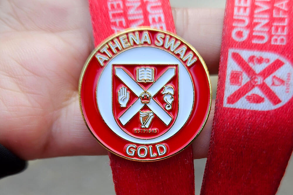 A close-up of a hand holding a red lanyard with a gold Athena SWAN Gold award pin featuring Queen's University Belfast insignia.